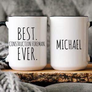 May include: Two white ceramic mugs with black handles and black rims. One mug says "Best. Construction Foreman. Ever." and the other mug says "Michael".