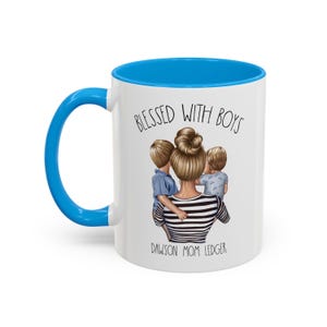 Puede incluir: Taza de cer&aacute;mica blanca con borde y asa azules. La taza presenta un gr&aacute;fico de una mujer con dos ni&ntilde;os, con el texto "Blessed with Boys" y "Dawson Mom Ledger".