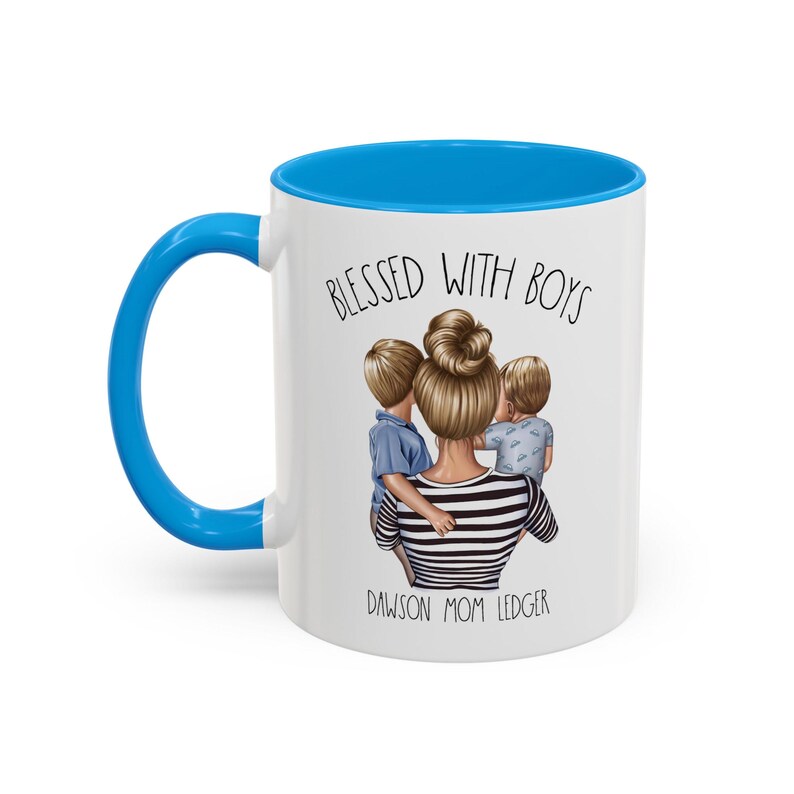Puede incluir: Taza de cer&aacute;mica blanca con borde y asa azules. La taza presenta un gr&aacute;fico de una mujer con dos ni&ntilde;os, con el texto "Blessed with Boys" y "Dawson Mom Ledger".