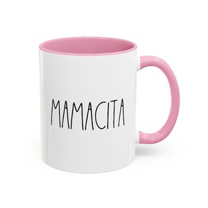 May include: White ceramic mug with a pink interior and handle. The word "MAMACITA" is printed in black, handwritten-style lettering on the front of the mug. The mug is designed for hot or cold beverages.