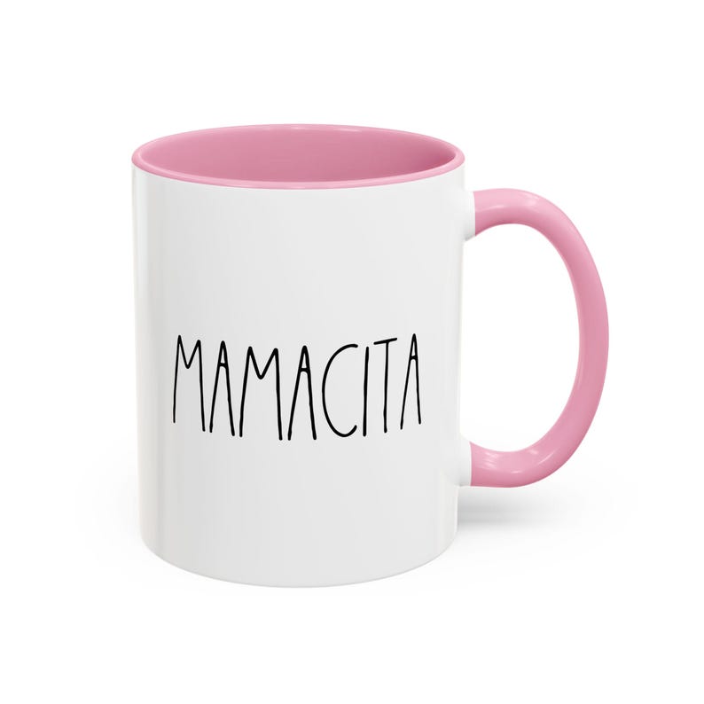 May include: White ceramic mug with a pink interior and handle. The word "MAMACITA" is printed in black, handwritten-style lettering on the front of the mug. The mug is designed for hot or cold beverages.