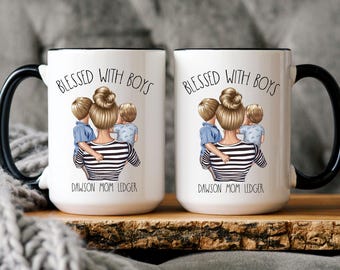 May include: Two white ceramic mugs with black handles and black rims. Both mugs feature a design of a woman with two children, with the text "Blessed with Boys" and the names "Dawson Mom Ledger".