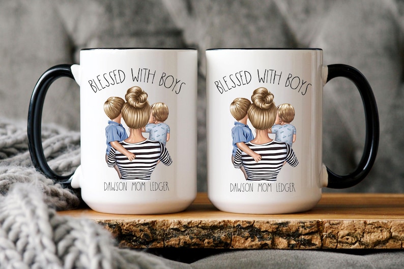 Puede incluir: Dos tazas de cer&aacute;mica blanca con asas negras y bordes negros. Ambas tazas presentan un dise&ntilde;o de una mujer con dos ni&ntilde;os, con el texto "Blessed with Boys" y los nombres "Dawson Mom Ledger".