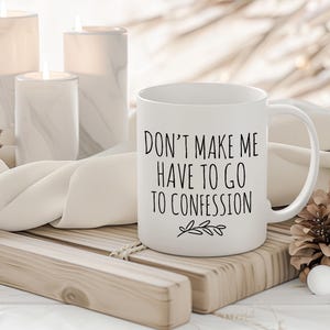 May include: White ceramic mug with the text "DON'T MAKE ME HAVE TO GO TO CONFESSION" in black script. The mug is on a wooden tray with candles and pinecones in the background. A decorative item for the home.