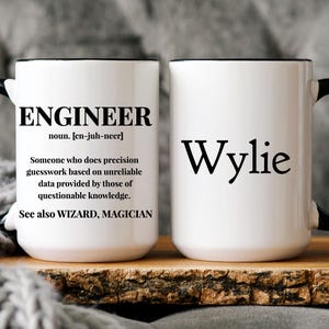 May include: Two white ceramic mugs with black handles. One mug has the definition of "engineer" printed on it. The other mug has the name "Wylie" printed on it.