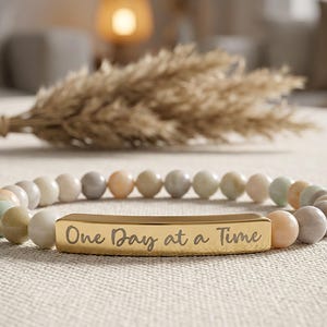 May include: A beaded bracelet with a gold bar engraved with the words "One Day at a Time." The bracelet features a variety of pastel-colored beads, including shades of green, peach, and gray. The bracelet is displayed on a neutral-colored surface.