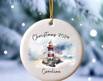 May include: White ceramic Christmas ornament with a watercolor painting of a red and white lighthouse on a rocky shore. The ornament has the text "Christmas 2024" and "Caroline" on it.