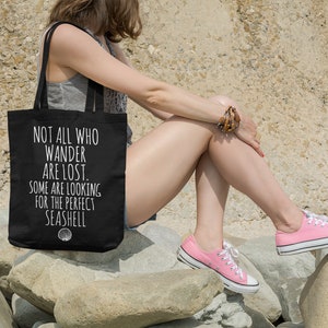 May include: A black tote bag with white text that reads "Not all who wander are lost. Some are looking for the perfect seashell."  A seashell icon is below the text.