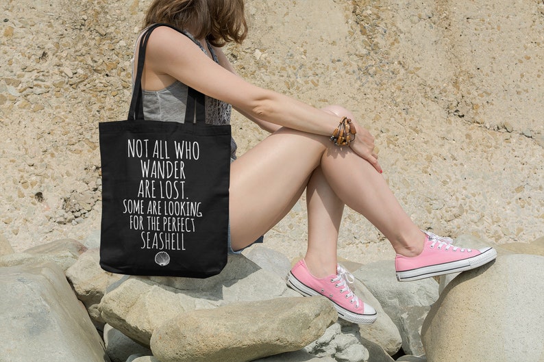 May include: A black tote bag with white text that reads "Not all who wander are lost. Some are looking for the perfect seashell."  A seashell icon is below the text.