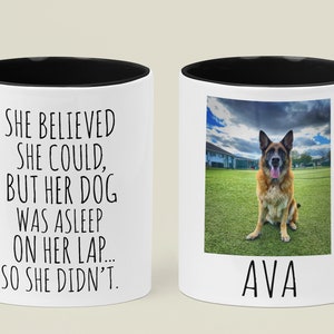 Op de afbeelding: Een witte keramische mok met een zwarte rand en handvat. De mok heeft een zwart-wit tekstontwerp met de tekst "Ze geloofde dat ze kon, maar haar hond sliep op haar schoot... dus deed ze het niet." De andere kant van de mok heeft een foto van een Duitse herder die op een grasveld zit met de naam "AVA" onder de foto.