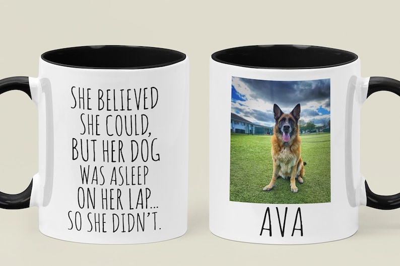 Op de afbeelding: Een witte keramische mok met een zwarte rand en handvat. De mok heeft een zwart-wit tekstontwerp met de tekst "Ze geloofde dat ze kon, maar haar hond sliep op haar schoot... dus deed ze het niet." De andere kant van de mok heeft een foto van een Duitse herder die op een grasveld zit met de naam "AVA" onder de foto.
