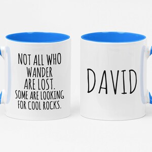 K&ouml;nnte beinhalten: Zwei wei&szlig;e Keramikbecher mit blauen R&auml;ndern und Griffen. Ein Becher hat den Text "NOT ALL WHO WANDER ARE LOST. SOME ARE LOOKING FOR COOL ROCKS." Der andere Becher hat den Namen "DAVID" aufgedruckt.