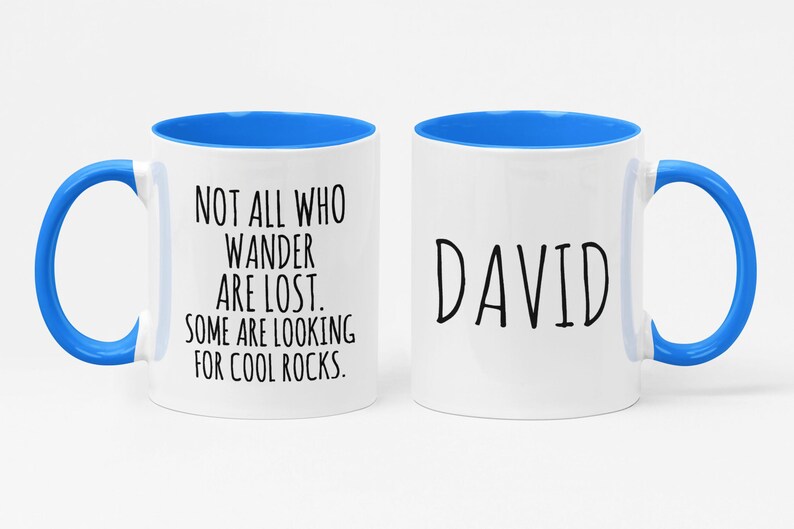 K&ouml;nnte beinhalten: Zwei wei&szlig;e Keramikbecher mit blauen R&auml;ndern und Griffen. Ein Becher hat den Text "NOT ALL WHO WANDER ARE LOST. SOME ARE LOOKING FOR COOL ROCKS." Der andere Becher hat den Namen "DAVID" aufgedruckt.