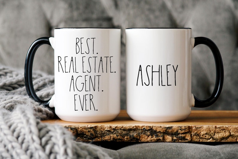 May include: Two white ceramic mugs with black handles. One mug says "Best. Real Estate. Agent. Ever." and the other mug says "Ashley".