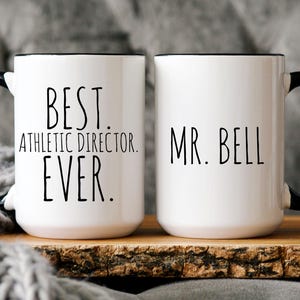 May include: Two white ceramic mugs with black handles and black rims. One mug says "Best Athletic Director Ever." The other mug says "Mr. Bell."