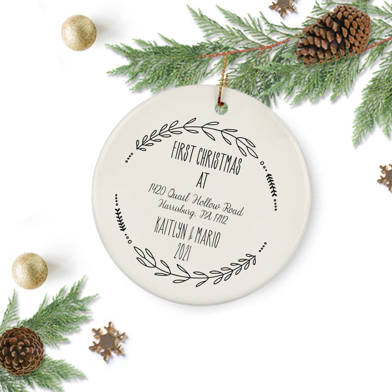 May include: White ceramic Christmas ornament with black text that reads "First Christmas at 1420 Quail Hollow Road Harrisburg, PA 17112 Kaitlyn & Mario 2021" surrounded by a wreath of black leaves.