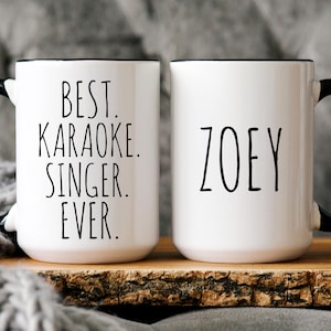 May include: Two white ceramic mugs with black handles and rims. One mug reads "BEST. KARAOKE. SINGER. EVER." in black lettering. The other mug has the name "ZOEY" in black lettering. Both mugs are on a wooden surface.