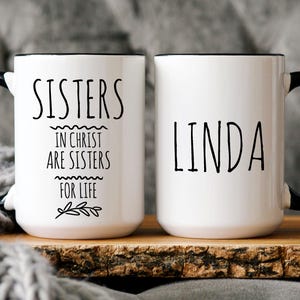 May include: Two white ceramic mugs with black handles and black rims. One mug says "Sisters in Christ are sisters for life" and the other mug says "Linda".