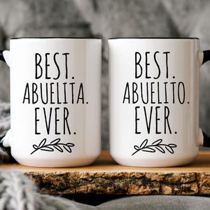 Peut inclure: Deux tasses en c&eacute;ramique blanche avec des poign&eacute;es noires et des bords noirs. La tasse de gauche dit "Best Abuelita Ever." La tasse de droite dit "Best Abuelito Ever."
