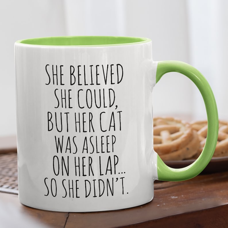 Puede incluir: Taza de caf&eacute; de cer&aacute;mica blanca con borde y asa verdes. La taza tiene texto negro que dice "SHE BELIEVED SHE COULD, BUT HER CAT WAS ASLEEP ON HER LAP... SO SHE DIDN'T."