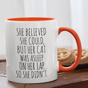Puede incluir: Taza de caf&eacute; de cer&aacute;mica blanca con borde y asa naranja. La taza tiene un dicho divertido impreso: "Ella cre&iacute;a que pod&iacute;a, pero su gato estaba dormido en su regazo... as&iacute; que no lo hizo."
