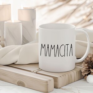 May include: White ceramic mug with the word "Mamacita" in black script. The mug is on a wooden tray with candles and pinecones in the background. The mug has a curved handle and a smooth surface.