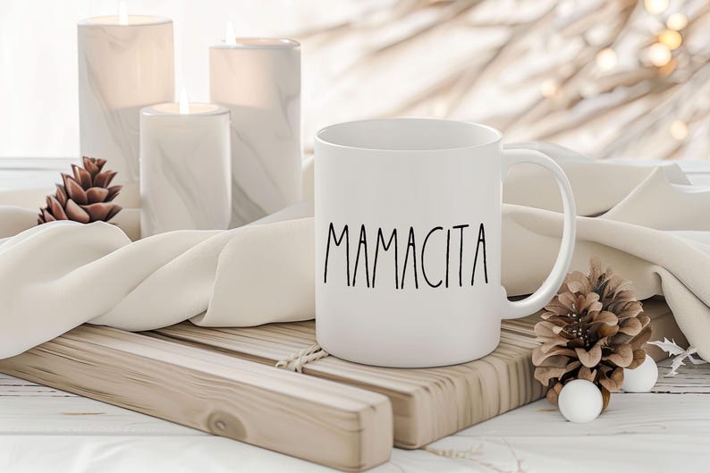 May include: White ceramic mug with the word "Mamacita" in black script. The mug is on a wooden tray with candles and pinecones in the background. The mug has a curved handle and a smooth surface.