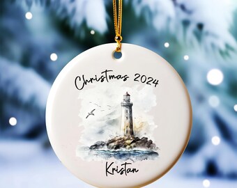 May include: Round white Christmas ornament with a watercolor painting of a lighthouse on a rocky shore. The text "Christmas 2024" and a name are written in black script. A gold string is attached for hanging.