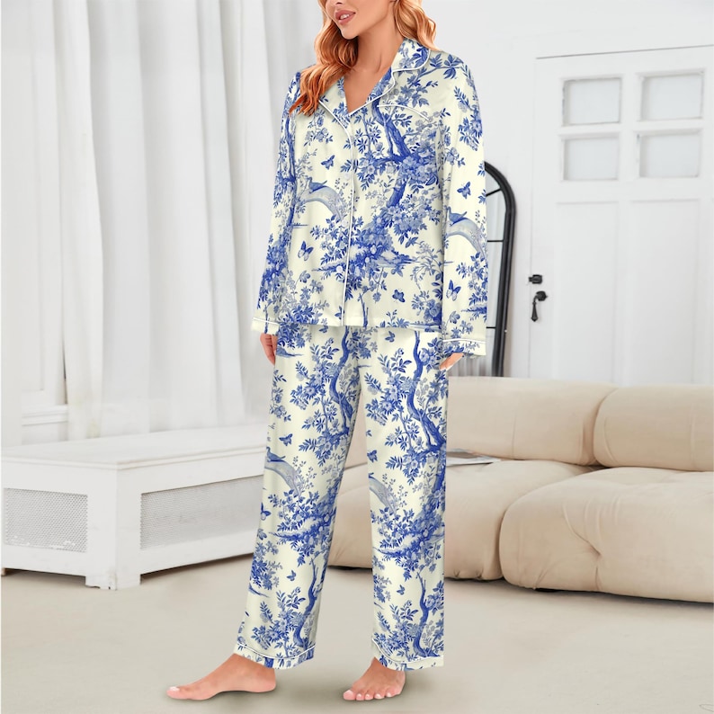 May include: Cream-coloured pyjama set with a blue toile pattern featuring trees, birds, and butterflies. The long-sleeved top has a collared neckline and button closure. The trousers are straight-legged. White trim accents the edges of the pyjamas.