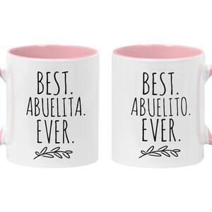 Peut inclure: Deux tasses en c&eacute;ramique blanche avec des bords et des poign&eacute;es roses. La tasse de gauche dit "Best Abuelita Ever." La tasse de droite dit "Best Abuelito Ever."