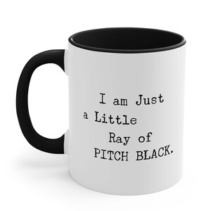 May include: White ceramic coffee mug with a black rim and handle. The mug has black text that reads "I am Just a Little Ray of PITCH BLACK."