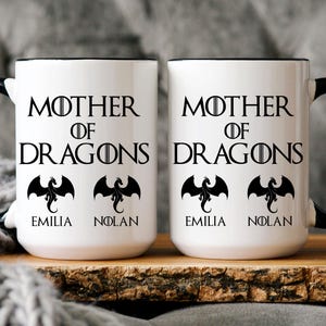 May include: Two white ceramic mugs with black handles and black rims. The mugs feature the text "Mother of Dragons" with a dragon graphic and the names "Emilia" and "Nolan" printed on them.