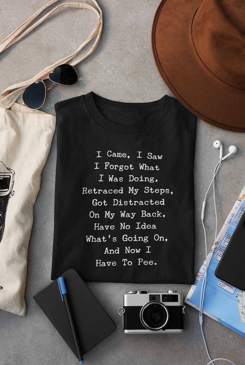 May include: Black t-shirt with white text: "I Came, I Saw, I Forgot What I Was Doing, Retraced My Steps, Got Distracted, On My Way Back, Have No Idea What's Going On, And Now I Have To Pee." Accessories include sunglasses, a tote bag, and a camera.
