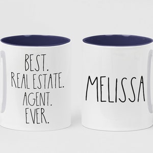 May include: Two white ceramic mugs with blue rims and handles. One mug says "BEST. REAL ESTATE. AGENT. EVER." and the other mug says "MELISSA".