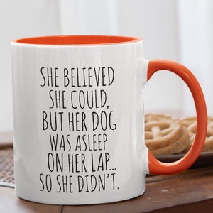 Op de afbeelding: Een witte koffiemok met een oranje rand en handvat. De mok heeft zwarte tekst met de tekst "SHE BELIEVED SHE COULD, BUT HER DOG WAS ASLEEP ON HER LAP... SO SHE DIDN'T."