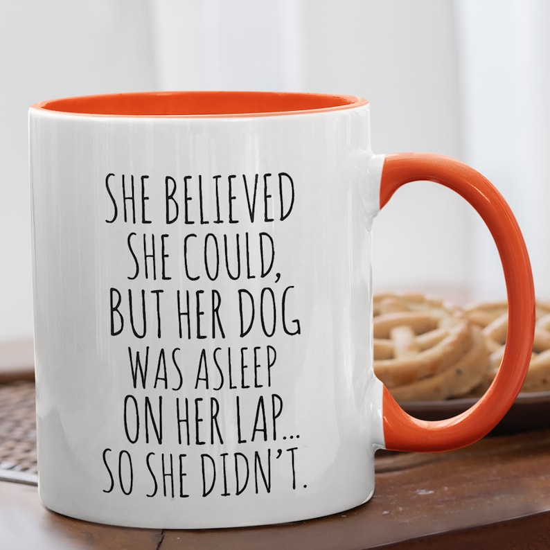 Op de afbeelding: Een witte koffiemok met een oranje rand en handvat. De mok heeft zwarte tekst met de tekst "SHE BELIEVED SHE COULD, BUT HER DOG WAS ASLEEP ON HER LAP... SO SHE DIDN'T."