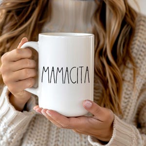 May include: White ceramic mug with the word "MAMACITA" printed in black, held by a person wearing a beige sweater. The mug has a large handle and a simple, clean design, suitable for hot beverages.