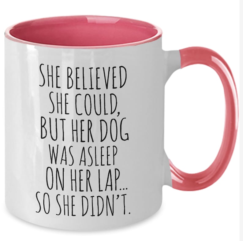 Op de afbeelding: Witte keramische koffiemok met een roze rand en handvat. De mok heeft een grappige spreuk erop gedrukt: "Ze geloofde dat ze kon, maar haar hond sliep op haar schoot... dus deed ze het niet."
