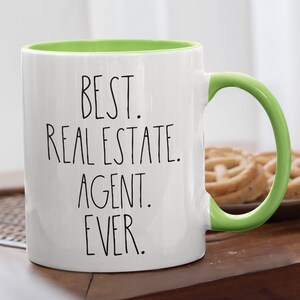 May include: White ceramic coffee mug with a green rim and handle. The mug has black text that reads "BEST. REAL ESTATE. AGENT. EVER."