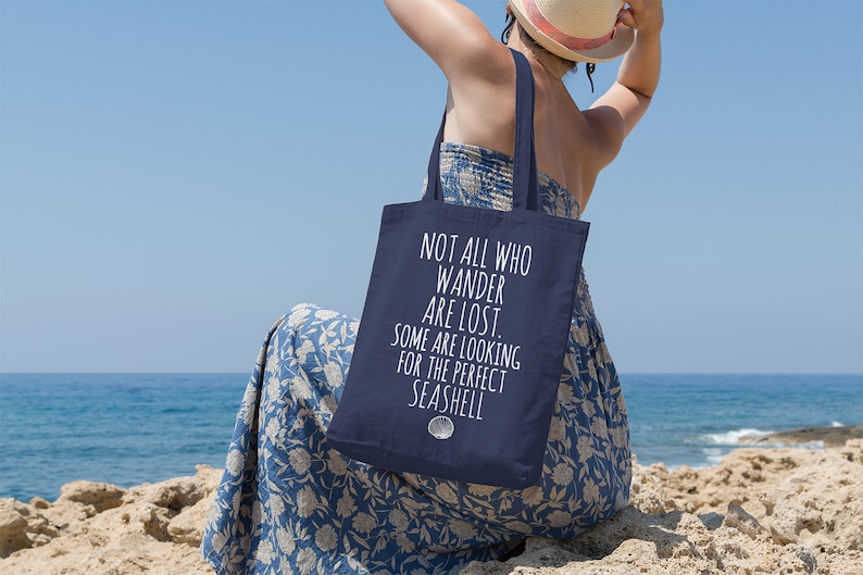May include: A navy blue tote bag with white text that reads "Not all who wander are lost. Some are looking for the perfect seashell." The bag is being carried by a person sitting on a rocky beach.