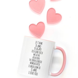 May include: White ceramic mug with a pink handle. The mug has black text that reads "Je t'aime, Te amo, Se agapo, Ich liebe dich, Wo ai ni, Aishiteru, Ya tebya liubliu, Nemehotatse, Is brea liom tu, Ti amo, I love you".