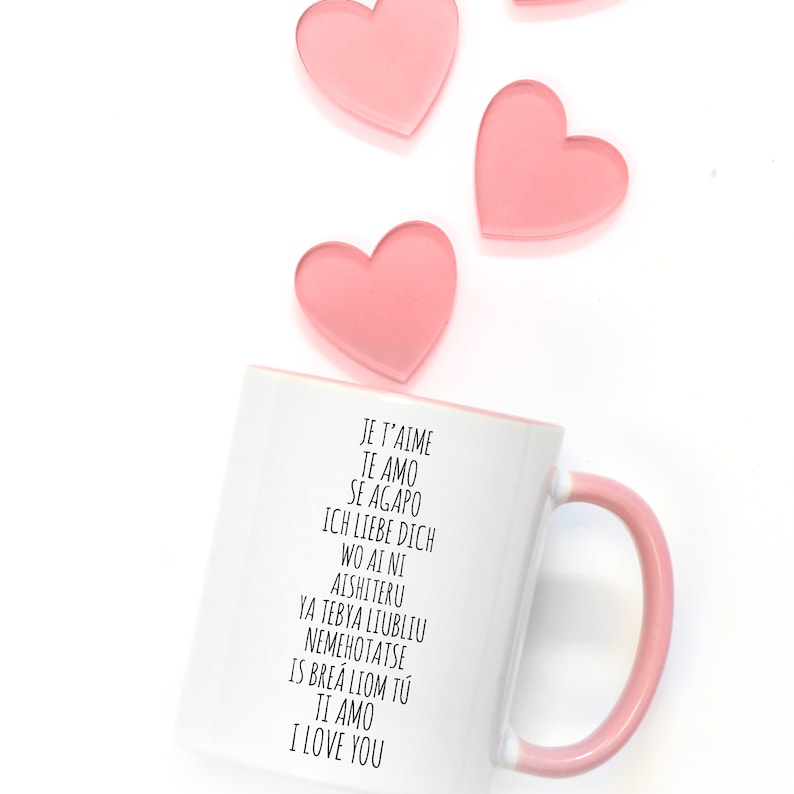 May include: White ceramic mug with a pink handle. The mug has black text that reads "Je t'aime, Te amo, Se agapo, Ich liebe dich, Wo ai ni, Aishiteru, Ya tebya liubliu, Nemehotatse, Is brea liom tu, Ti amo, I love you".