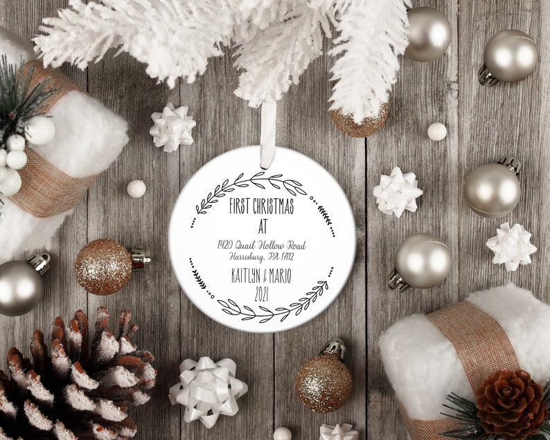 May include: White round Christmas ornament with black text that reads "First Christmas at 1420 Quail Hollow Road Harrisburg, PA 17112 Kaitlyn & Mario 2021". The ornament is surrounded by a wreath of leaves.