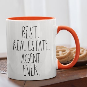 May include: White ceramic coffee mug with an orange rim and handle. The mug has black text that reads "BEST. REAL ESTATE. AGENT. EVER."