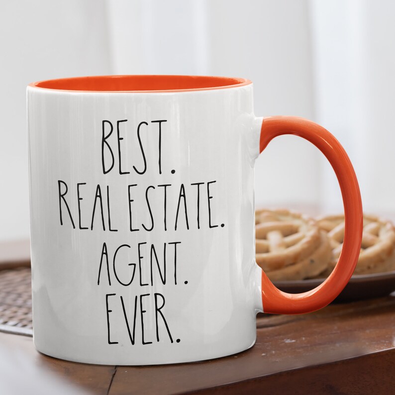 May include: White ceramic coffee mug with an orange rim and handle. The mug has black text that reads "BEST. REAL ESTATE. AGENT. EVER."