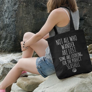 May include: A black tote bag with white text that reads "Not all who wander are lost. Some are looking for the perfect seashell."  The bag is sitting on a rock by the ocean.