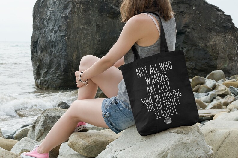 May include: A black tote bag with white text that reads "Not all who wander are lost. Some are looking for the perfect seashell."  The bag is sitting on a rock by the ocean.