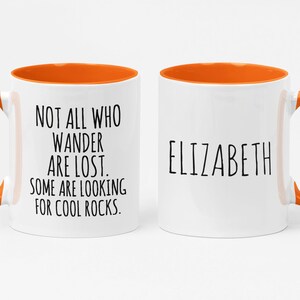 K&ouml;nnte beinhalten: Zwei wei&szlig;e Keramikbecher mit orangefarbenen R&auml;ndern und Griffen. Ein Becher tr&auml;gt den Text "NOT ALL WHO WANDER ARE LOST. SOME ARE LOOKING FOR COOL ROCKS." Der andere Becher tr&auml;gt den Namen "ELIZABETH" aufgedruckt.