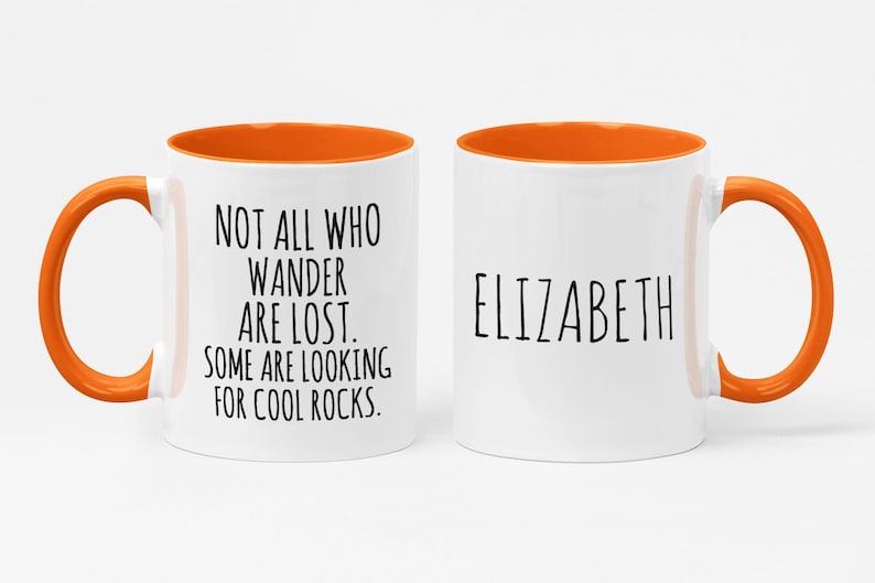 K&ouml;nnte beinhalten: Zwei wei&szlig;e Keramikbecher mit orangefarbenen R&auml;ndern und Griffen. Ein Becher tr&auml;gt den Text "NOT ALL WHO WANDER ARE LOST. SOME ARE LOOKING FOR COOL ROCKS." Der andere Becher tr&auml;gt den Namen "ELIZABETH" aufgedruckt.