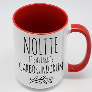 May include: White ceramic coffee mug with a red rim and handle. The mug has black text that reads "Nolite Te Bastardes Carborundum" with a small branch design below the text.